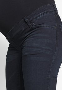 Jeans de maternidade em denim azul escuro com uma textura suave, cintura média e painéis laterais para conforto. Apresenta costuras clássicas e bolsos.