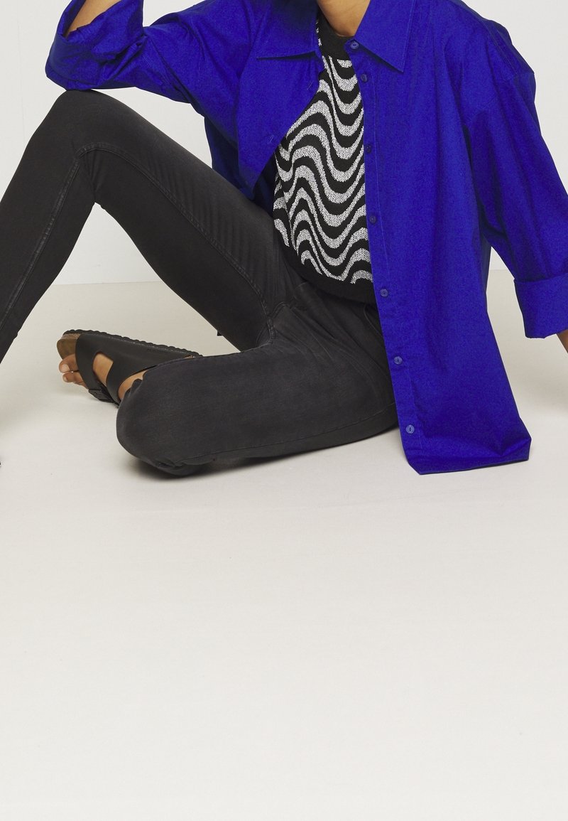 Camisa de gran tamaño azul brillante, top negro con estampado, jeans ajustados negros y sandalias negras, con una textura suave y un sutil patrón de onda en la parte superior.