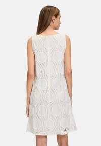 Weißes ärmelloses Kleid mit einer strukturierten Spitzenoberfläche mit kreisförmigen Mustern, die ein leichtes und luftiges Erscheinungsbild verleihen.