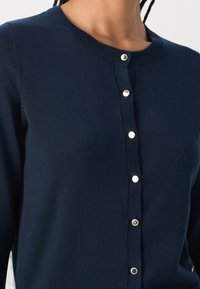 Námořnická modrá pletená bunda s kulatým výstřihem a stříbrnými cvoky vpředu, kterou nosí osoba s copy.