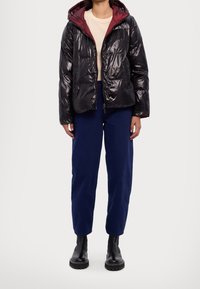 Veste matelassée noire à finition brillante, doublure intérieure bordeaux, fermeture éclair frontale et capuche. Assortie à un pantalon bleu marine à coupe décontractée et des bottes noires.