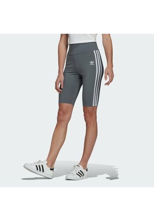 Osoba ubrana w szare spodenki rowerowe Adidas z białymi paskami i białe buty sportowe Adidas, stojąca na jednej nodze na białym tle.
