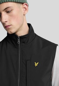 Schwarze ärmellose Jacke mit einem Reißverschluss vorne, zwei Taschen und einem gelben, gestickten Logo auf der linken Brust. Glatte Textur.
