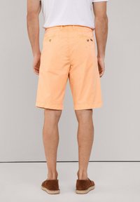 Pantalones cortos naranja claro con un frente plano, bolsillos traseros con botones y un ajuste entallado. Se llevan con alpargatas marrones.