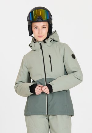 Jonge vrouw in een lichtgroene en grijze skijack met capuchon, met een zwarte skihelm en reflecterende goggles, staande tegen een witte achtergrond.