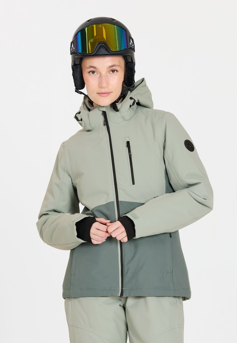 Giovane donna in giacca da sci leggera verde chiaro e grigia con cappuccio, che indossa un casco da sci nero e occhiali da sci riflettenti, in piedi su sfondo bianco.