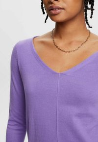 Fioletowy sweter z długim rękawem, zapinany na V, z delikatnym splotem. Posiada centralny szew i jest noszony z naszyjnikiem w złotym kolorze.