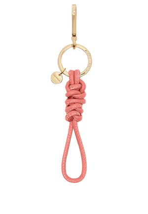 Portachiavi in pelle di un delicato rosa, caratterizzato da un design annodato, un anello in metallo dorato e un piccolo ciondolo inciso attaccato. Texture liscia.