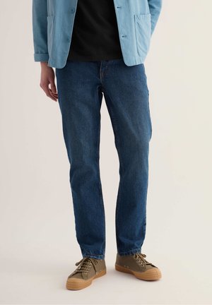 Mann trägt blaue Jeans, olivgrüne Sneakers mit Gummisohlen, ein schwarzes Hemd und eine hellblaue Freizeitjacke und steht vor einem hellen Hintergrund.