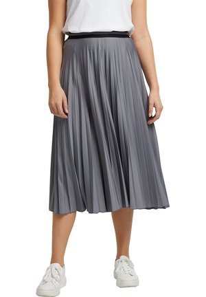 Femme portant une jupe midi plissée grise, un t-shirt blanc rentré à l'intérieur, et des baskets blanches, debout les bras détendus le long du corps.