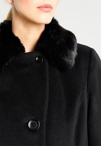 Zwarte wollen jas met een luxe zwart nepbont kraag, twee knopen en een getailleerd silhouet. Glad textuur met strakke lijnen.