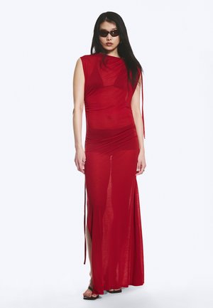 Femme portant une robe longue rouge sans manches et transparente, avec des fentes latérales et des sandales noires, posant devant un fond blanc uni.
