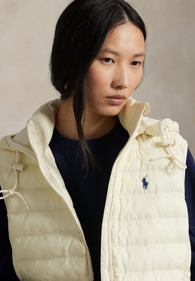 Kvinne med langt svart hår iført en kremfarget boblevest over en mørk topp, som ser nedover mot en nøytral bakgrunn.