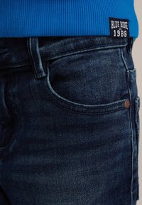 Tumši zili denim džinsi ar izbalējušu tekstūru, ar priekšējo kabatu un birku, kas vēsta "BLUE RIDGE EST. 1986" uz zila džempera.