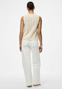 Krämfärgad ärmlös stickad topp med ett vertikalt mönster, i kombination med vita avslappnade byxor och slip-onsandaler.
