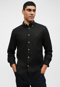 Čierna košeľa na gombíky s límcom, s jemným logom na hrudníku, v kombinácii s tmavými džínami. Hladká tkanina a priliehavý strih.