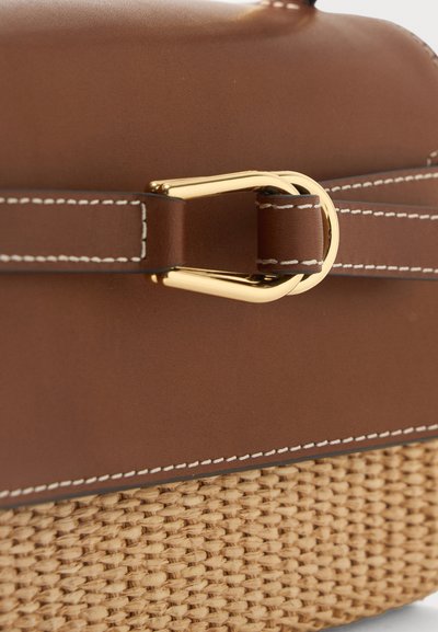 Καφέ δερμάτινη τσάντα με λευκά ραφές, χρυσή αγκράφα και λεπτομέρεια πλεγμένου καλαθιού στο κάτω μέρος σε κοντινή προβολή.