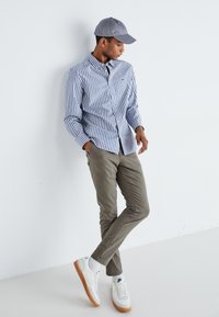Camicia a righe blu e bianche con bottoni, pantaloni kaki e sneakers bianche con suola in gomma. Un cappellino grigio completa il look.
