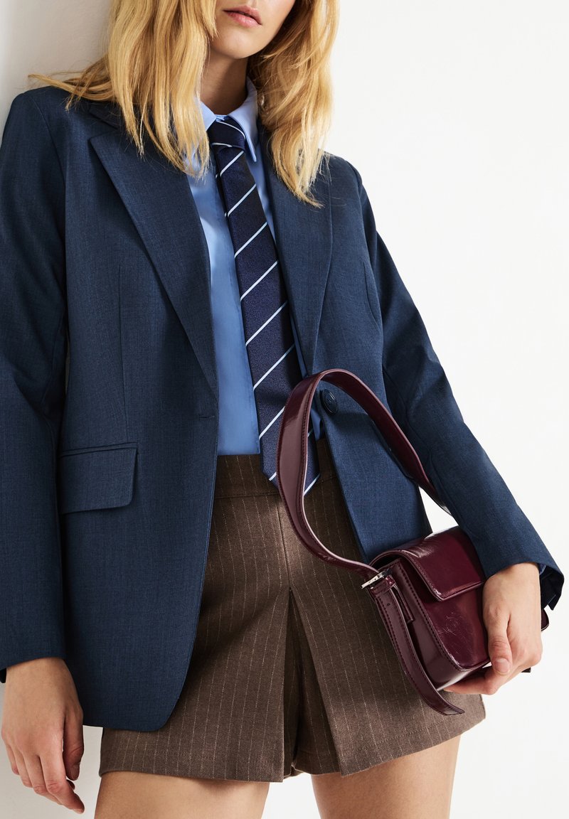 Donna bionda che indossa un blazer blu scuro, una camicia blu, una cravatta a righe, pantaloni corti a pieghe con righe marroni, mentre tiene una borsa in pelle rossa scura.