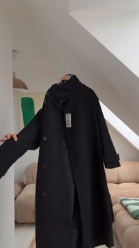 Svart kappa med hög krage, stora knappar framtill och långa ärmar. Hängande på en krok, texturerat tyg och en vid, svängd form.