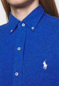 Blauwe polo met een gestructureerde brei, knoopsluiting, witte knopen en een klein geborduurd wit logo van een paard op de linkerkant van de borst.