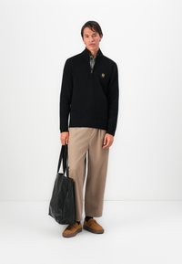 Homem em suéter preto de meia zip, calças largas bege, sapatos castanhos, segurando uma mala de mão de pele preta, em pé contra um fundo branco.