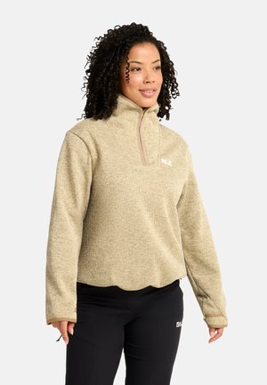 Donna con capelli ricci che indossa un pullover beige in pile con collo alto e pantaloni neri, in piedi davanti a uno sfondo semplice.