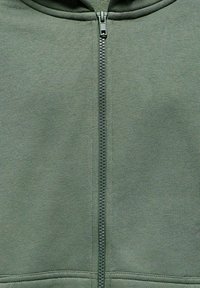 Hoodie zippé vert olive en tissu doux, avec une fermeture éclair à l'avant, des ourlets à double couture et une capuche ajustée.