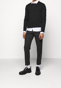 Pull en maille noire porté sur une chemise blanche à col, associé à un pantalon gris foncé et des chaussures noires. Design moderne avec coupe ajustée.