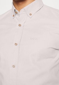 Camicia button-up grigia chiara con colletto a punta, caratterizzata da bottoni beige a contrasto e un logo in rilievo sul petto. Texture liscia.