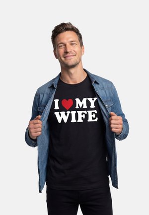 Een glimlachende man trekt zijn open denimshirt terug om een zwart T-shirt met witte tekst en een rood hart te onthullen met de tekst "Ik hou van mijn vrouw."