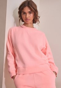 Light pink sweatshirt with textured fabric, oversized fit, and ribbed cuffs. It features a crew neckline and side pockets.