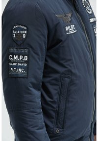 Marineblaue Bomberjacke mit gestickten Patches, Reißverschluss, seitlichen Taschen und einer glatten, leichten Stoffqualität.