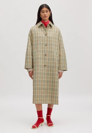 Femme debout portant un long manteau beige à carreaux sur un pull roulé rouge, avec des chaussettes rouges et des talons ouverts beiges sur un fond uni.