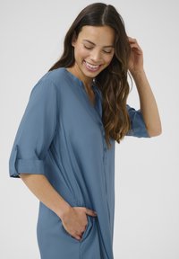 Abito camicia blu a maniche lunghe con pattina a bottoni, vestibilità comoda, tasche laterali e maniche arrotolate, realizzato in un tessuto leggero.