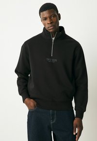 Black zip-neck sweatshirt made of soft material features a small embroidered "BROOKLYN" logo on the chest. Paired with dark denim pants.