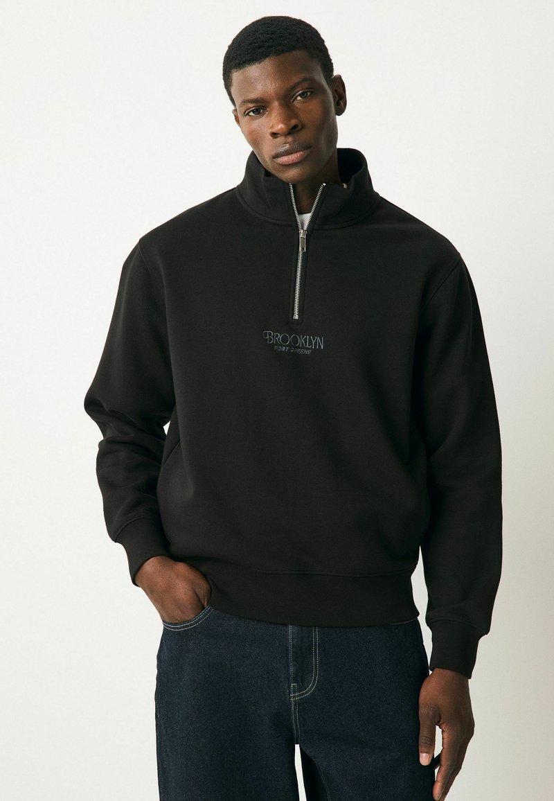 Svart zip-neck sweatshirt i mjukt material med en liten broderad "BROOKLYN"-logotyp på bröstet. Matchas med mörka jeans.
