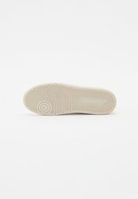 Semelle de sneaker blanche avec un motif en chevrons texturé, comportant un logo circulaire, des bords lisses et d'une couleur beige clair.