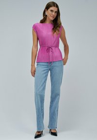 Mujer vistiendo una blusa sin mangas de color rosa con cinturilla, jeans de mezclilla azul claro, tacones negros y aretes de plata, de pie ante un fondo liso.