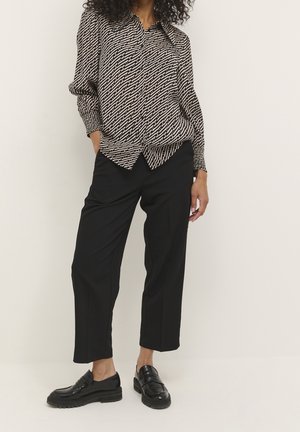 Femme portant un pantalon court noir, des mocassins noirs et un chemisier à manches longues à motif géométrique.