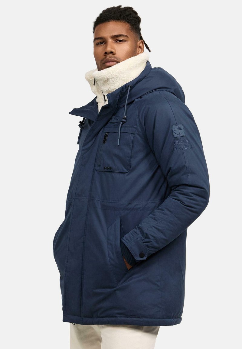 Mörkblå parka jacka med en krage i fuskpäls, flera fickor och justerbara snoddar. Slät tygstruktur, avslappnad passform.