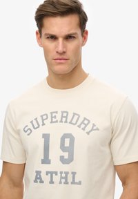 Camiseta color crema hecha de algodón con escote redondo. Presenta texto impreso en gris: "SUPERDRY 19 ATHL" centrado en la parte frontal.