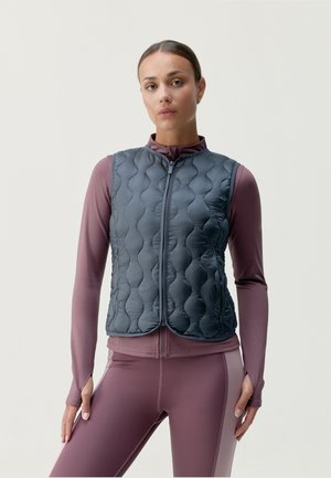 Gilet grigio scuro imbottito con motivo trapuntato, zip frontale completa e colletto alto, indossato sopra una maglietta a maniche lunghe di colore lilla chiaro.