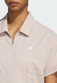 Ljusrosa sportpolotröja med korta ärmar, strukturerat randigt mönster, dragkedja framtill och en liten vit Adidas-logotyp på bröstet.