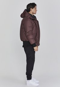 Maronfarbener Pufferjacke mit abnehmbarem Kapuzen, Frontreißverschluss und elastischen Bündchen, kombiniert mit schwarzen Cargo-Hosen und weißen Sneakern.