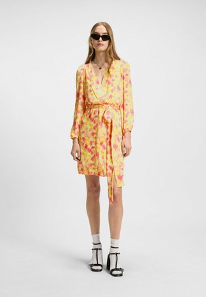 Floral wrap dress featuring yellow, pink, and orange hues, long sleeves, a tie waist, and a knee-length hem. Paired with black accessories.