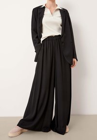 Černý tailored blazer přes bílý žebrovaný top, doplněný černými širokými kalhotami a světle růžovými teniskami, zdůrazňující štíhlý, minimalistick�ý design.