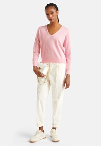 Mujer con suéter rosa de cuello en V y pantalones blancos sosteniendo un pequeño bolso blanco con cadena dorada, usando zapatillas blancas frente a un fondo blanco.