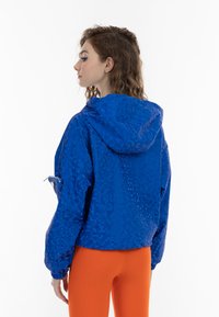 Giacca blu con cappuccio, caratterizzata da un motivo pixelato testurizzato, polsini elasticizzati e una tasca con zip sulla manica. Indossata sopra leggings arancioni brillanti.
