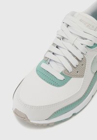 Tenisky Nike s bielym koženým povrchom, tyrkysovými akcentmi a svetlošedými detailmi. Majú vypchávku na jazyku, ploché šnúrky a textúrovanú podrážku.